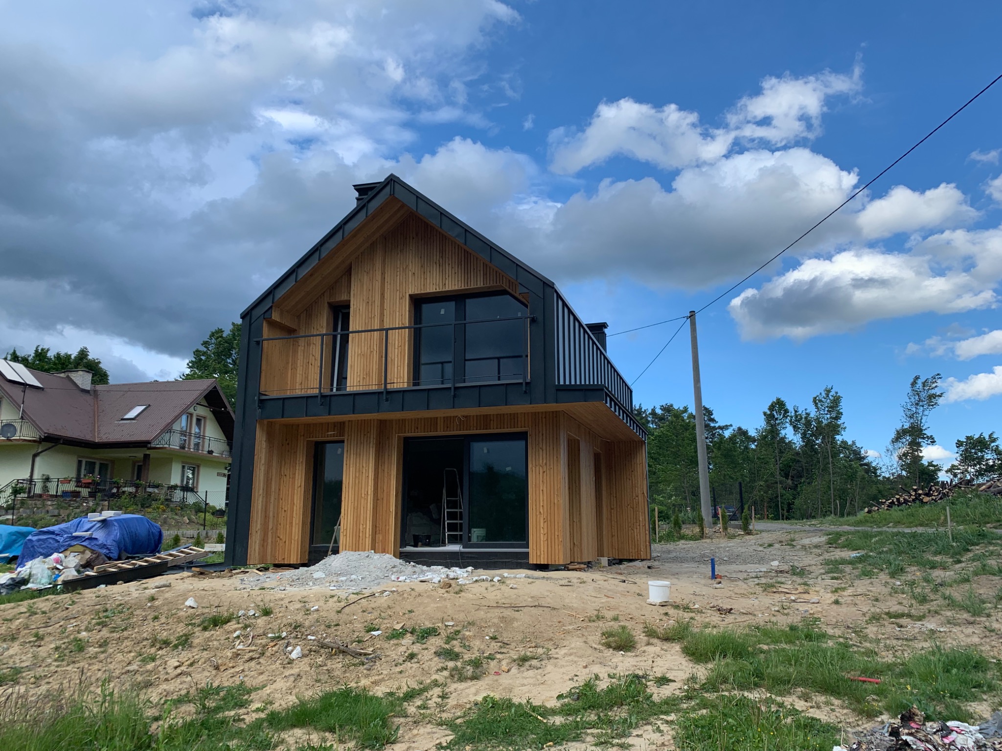Nowoczesny dom z drewnianą elewacją, czarnym dachem i balkonem, w trakcie budowy, widoczna drabina wewnątrz budynku, na pierwszym planie gruz i piasek.