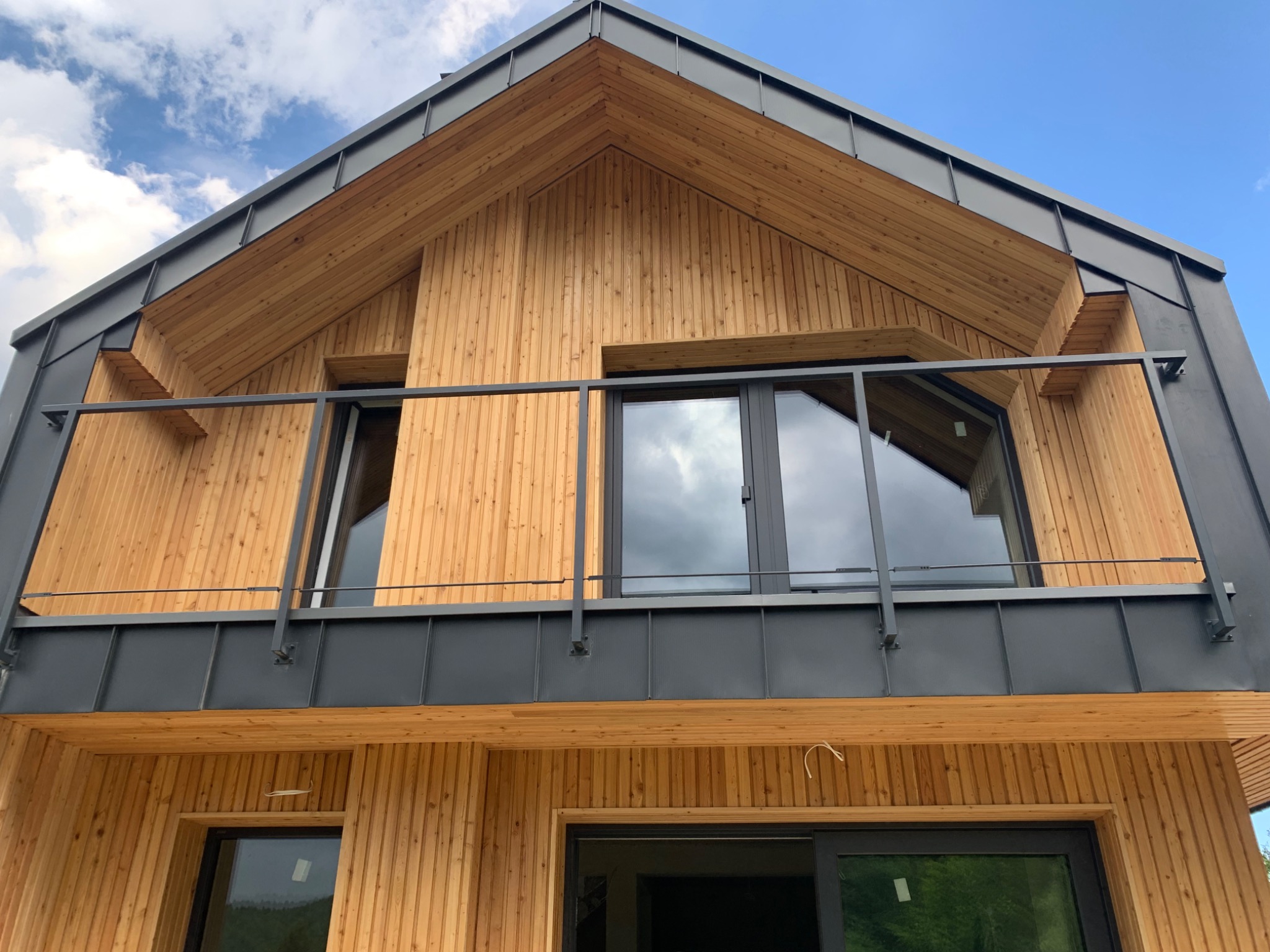 Fragment elewacji domu z drewnianą fasadą i czarnym dachem blaszanym, widok z dołu na okna i balkon z metalową balustradą.