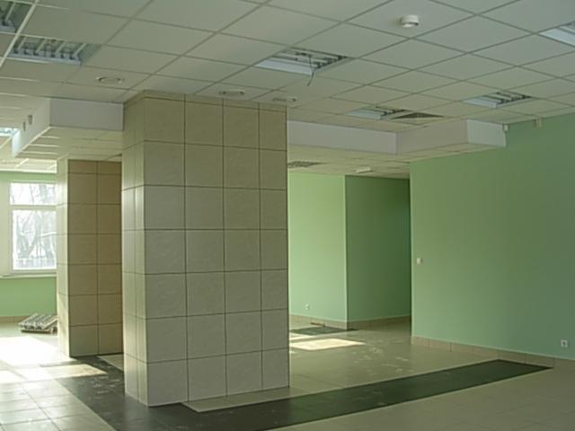 Wnętrze komercyjne z kolumnami wyłożonymi jasnymi płytkami ceramicznymi, zielonymi ścianami i podwieszanym sufitem kasetonowym.