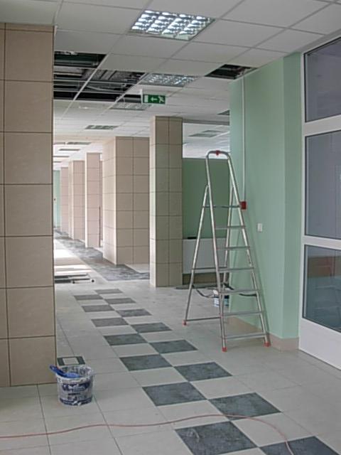 Korytarz w trakcie remontu z częściowo odsłoniętym sufitem podwieszanym, szachownicą na podłodze i aluminiową drabiną opartą o świeżo pomalowaną ścianę w odcieniu zieleni. Widoczny kubeł z farbą...