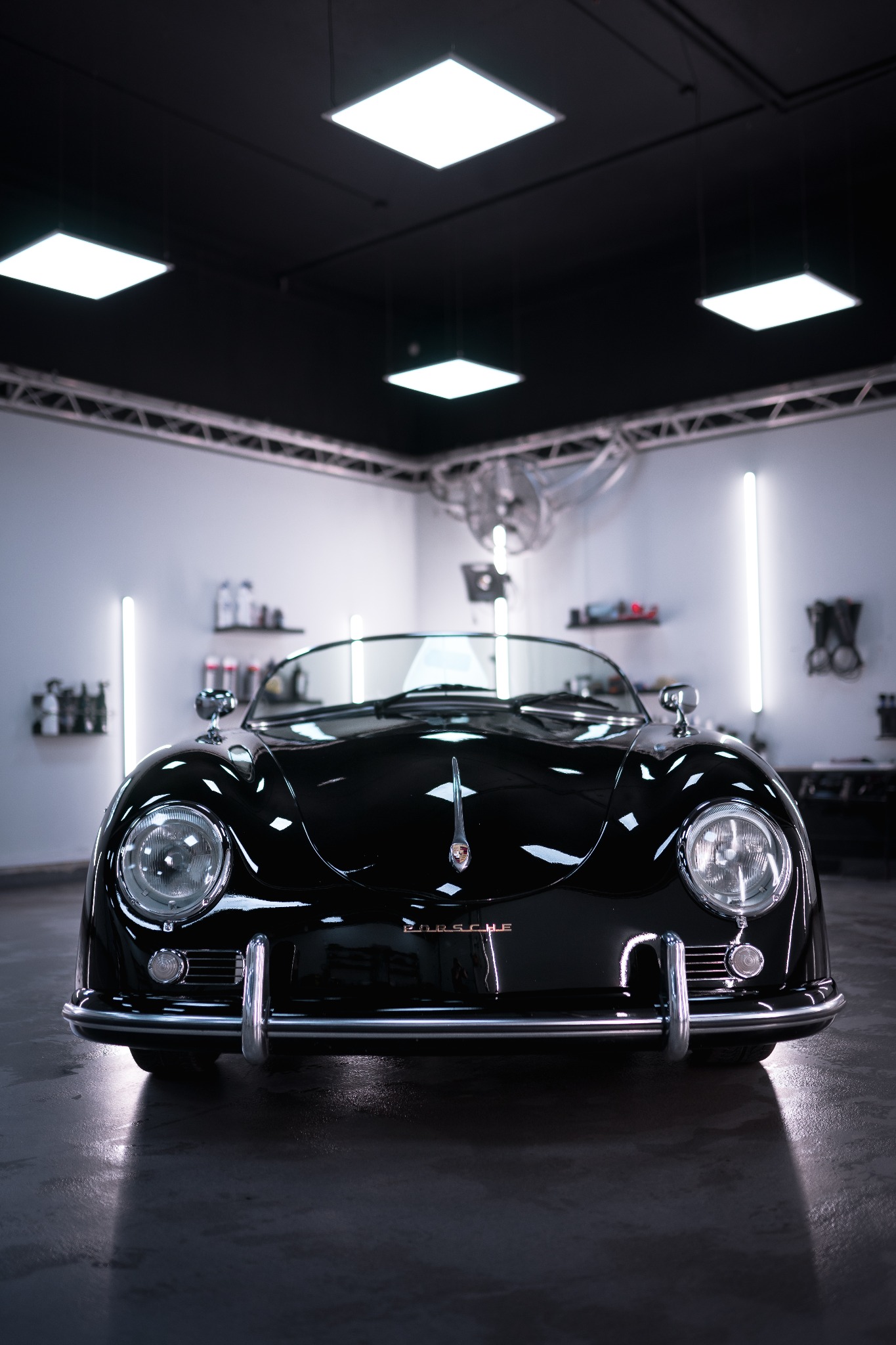 Przód odrestaurowanego czarnego Porsche Speedster z chromowanymi detalami, widoczny w jasnym warsztacie z oświetleniem LED, butelki z preparatami na półkach w tle.