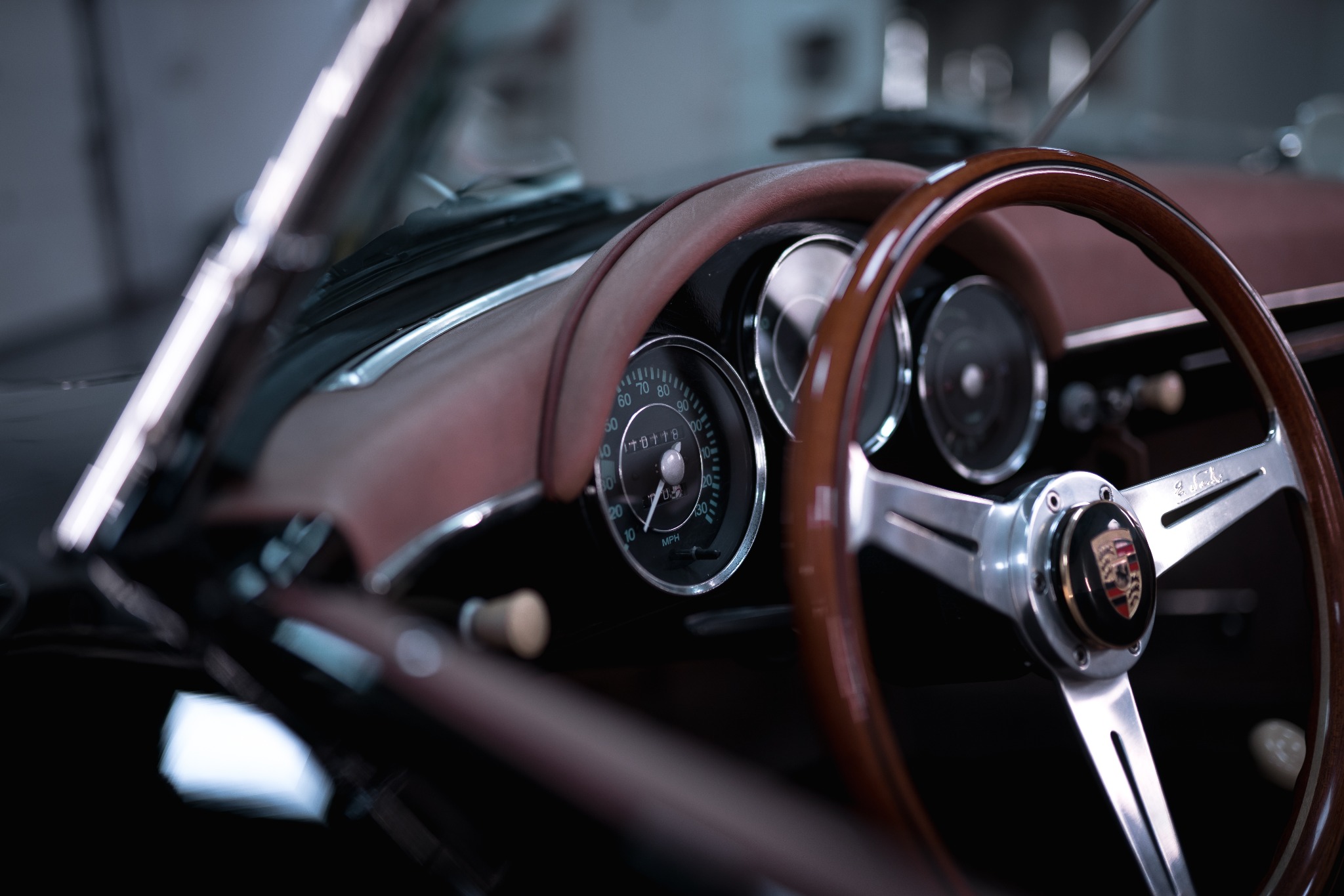 Wnętrze klasycznego Porsche Speedster z drewnianą kierownicą, chromowanymi elementami i brązową deską rozdzielczą; widoczny prędkościomierz i emblemat Porsche na kierownicy.