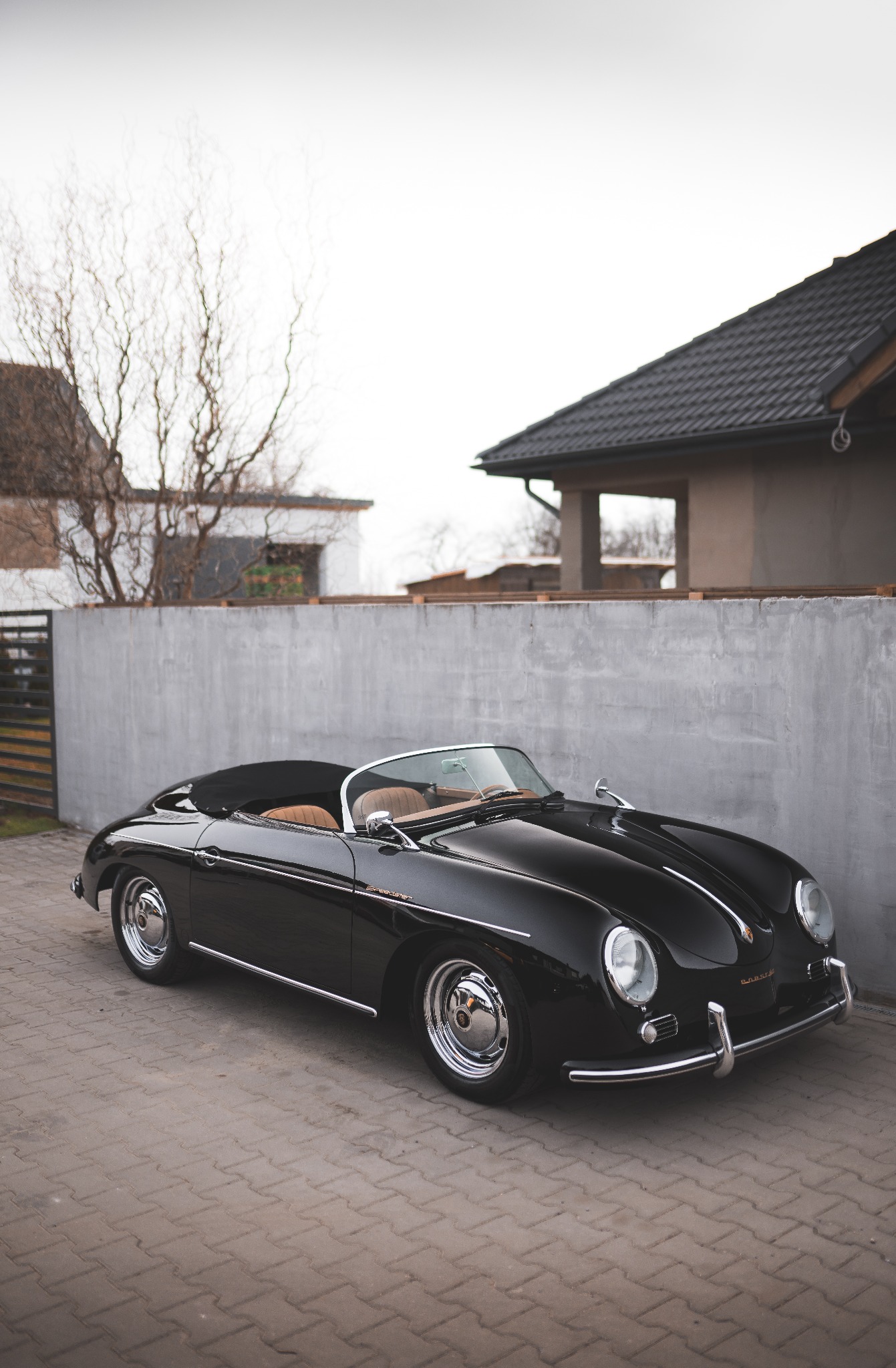 Czarny, klasyczny Porsche Speedster zaparkowany na brukowanej powierzchni, z szarą ścianą i domem w tle, prezentujący odrestaurowany stan.