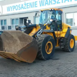 Żółta ładowarka kołowa marki ICE stoi na placu przed budynkiem 'Wyposażenie Warsztatów', w tle zaparkowane samochody.