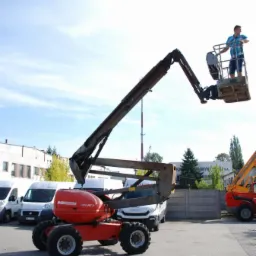 Czerwony podnośnik koszowy Maniaccess 160 ATJ z operatorem w koszu, na tle zaparkowanych białych samochodów dostawczych w Skwierzynie.