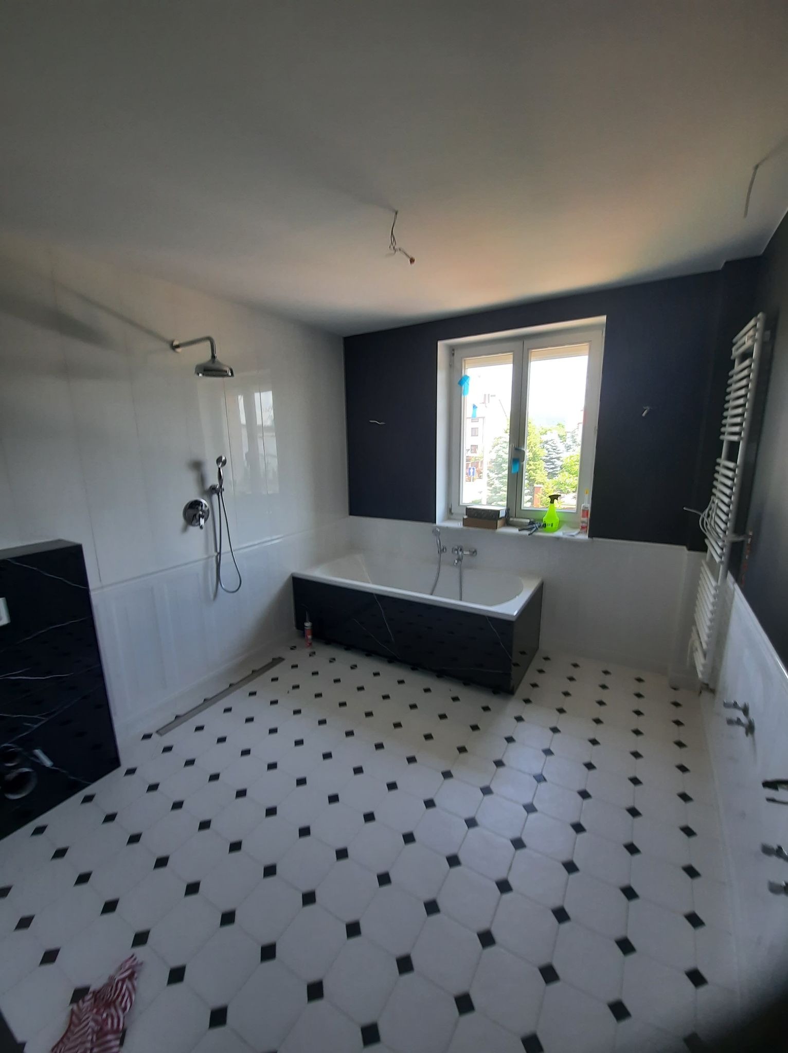 Wykończona łazienka z wanną obudowaną czarnym marmurem, biało-czarną mozaikową podłogą, chromowaną armaturą i grzejnikiem drabinkowym na tle granatowej ściany z oknem.