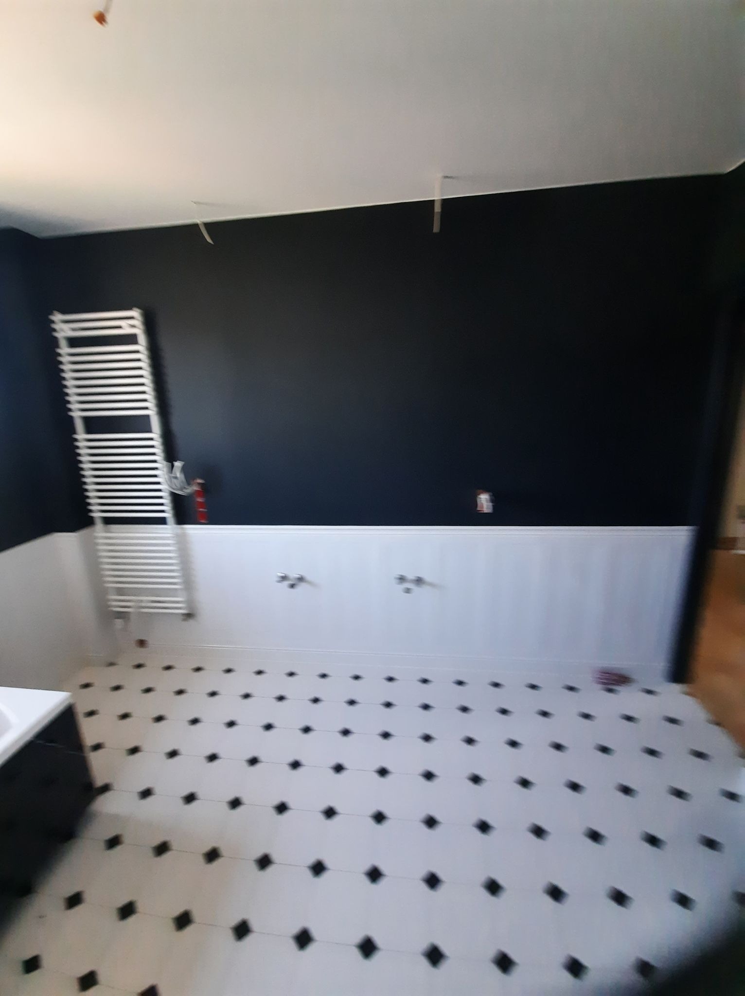 Częściowo wyremontowana łazienka z czarną ścianą, białymi panelami boazeryjnymi, grzejnikiem drabinkowym i biało-czarną podłogą w romby. Widoczne niedokończone prace przy suficie.