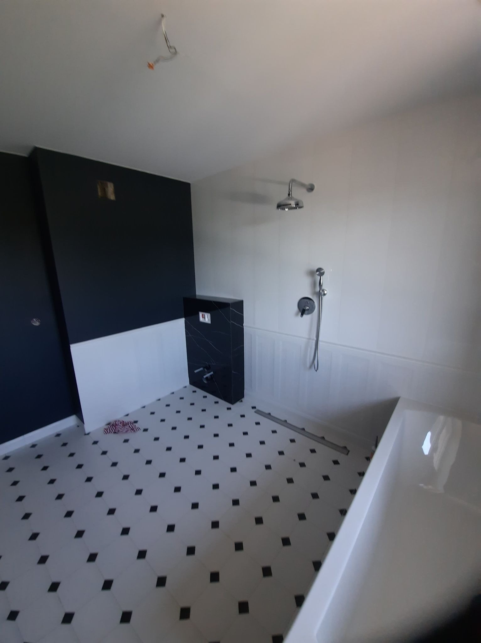 Wnętrze łazienki w trakcie remontu: czarno-biała mozaika na podłodze, częściowo wykończona ściana w kolorze granatowym i białym, zamontowana armatura prysznicowa, widoczne przewody elektryczne...