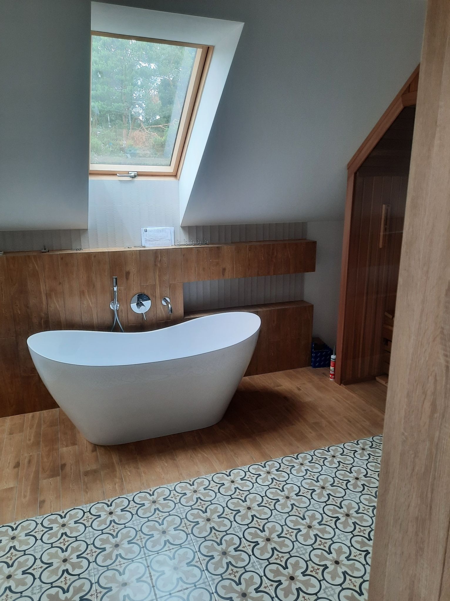 Nowoczesna łazienka na poddaszu z wolnostojącą białą wanną, drewnianą zabudową, wzorzystą podłogą i oknem dachowym.