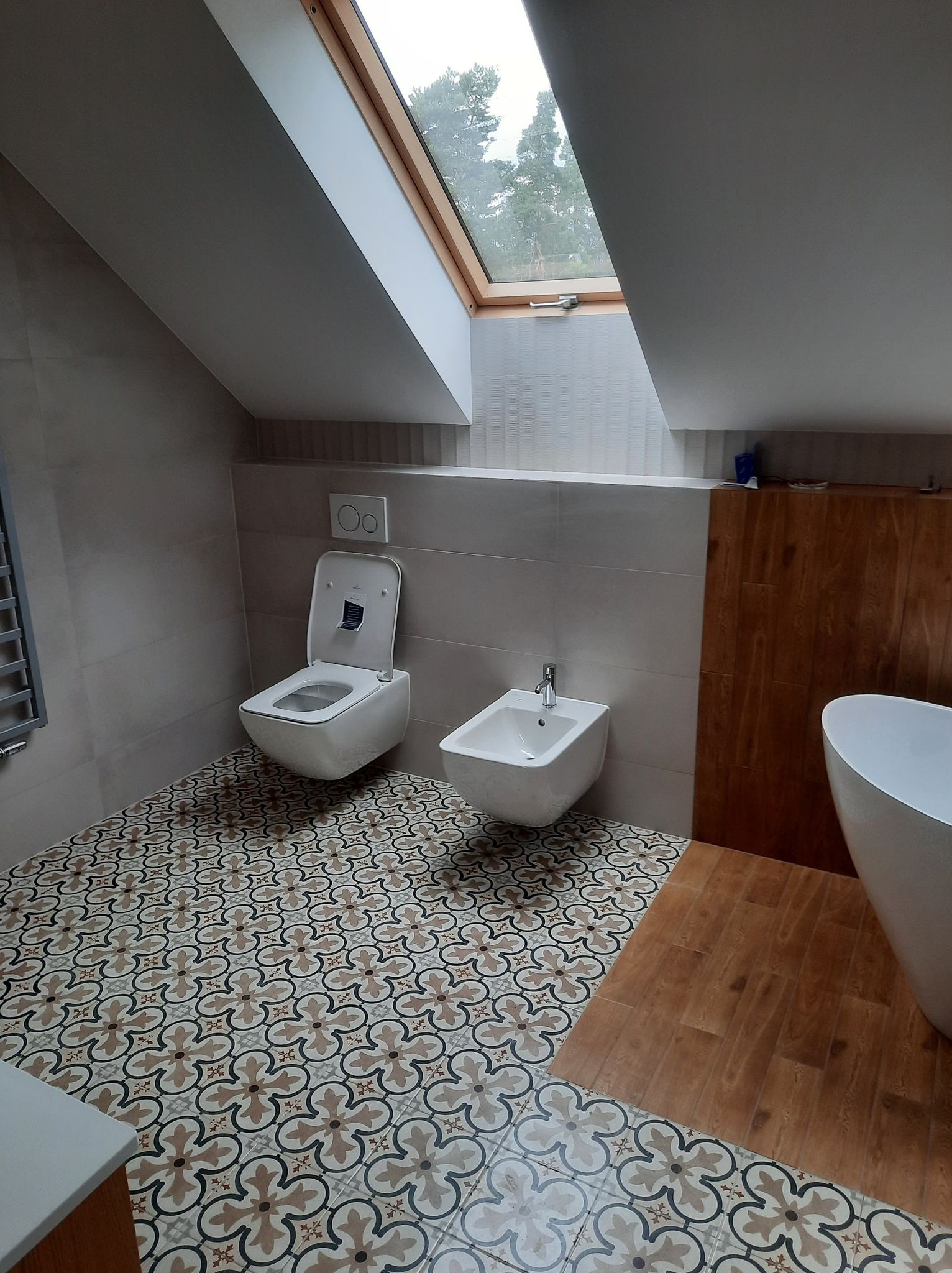 Wykończona łazienka na poddaszu ze skośnym oknem dachowym, z toaletą, bidetem i częścią wanny. Na podłodze wzorzyste płytki w stylu retro i fragment drewnianej podłogi.