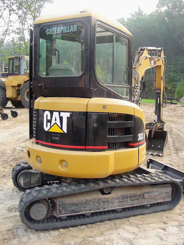 Żółto-czarna minikoparka Caterpillar 303 na gąsienicach, widoczna z przodu kabina z naklejką Caterpillar, w tle inna maszyna budowlana i drzewa.