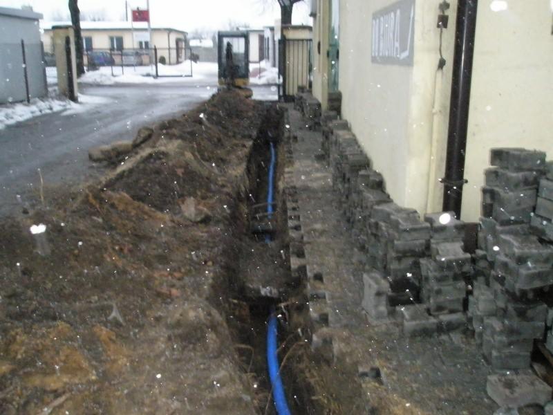 Wykop w ziemi z ułożonym niebieskim rurociągiem, częściowo zasypany, obok kostka brukowa i budynek w zimowej scenerii.