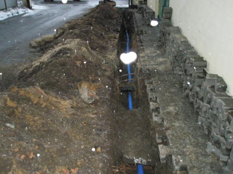 Wykop z ułożoną niebieską rurą wodociągową, widoczne fragmenty kostki brukowej i ziemi.