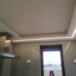 Nowoczesna kuchnia z podwieszanym sufitem i liniowym oświetleniem LED, widoczny okap ze stali nierdzewnej i okno z widokiem na zewnątrz.