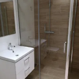 Nowoczesna łazienka z kabiną prysznicową typu walk-in z deszczownicą, umywalką nablatową na białej szafce z dwoma szufladami i lustrem. Płytki imitujące drewno na ścianach i podłodze.