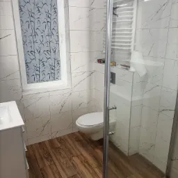 Nowoczesna łazienka po remoncie: białe marmurowe płytki ścienne, drewnopodobna podłoga, kabina prysznicowa ze szklanymi drzwiami, toaleta podwieszana i okno z folią imitującą bambus.