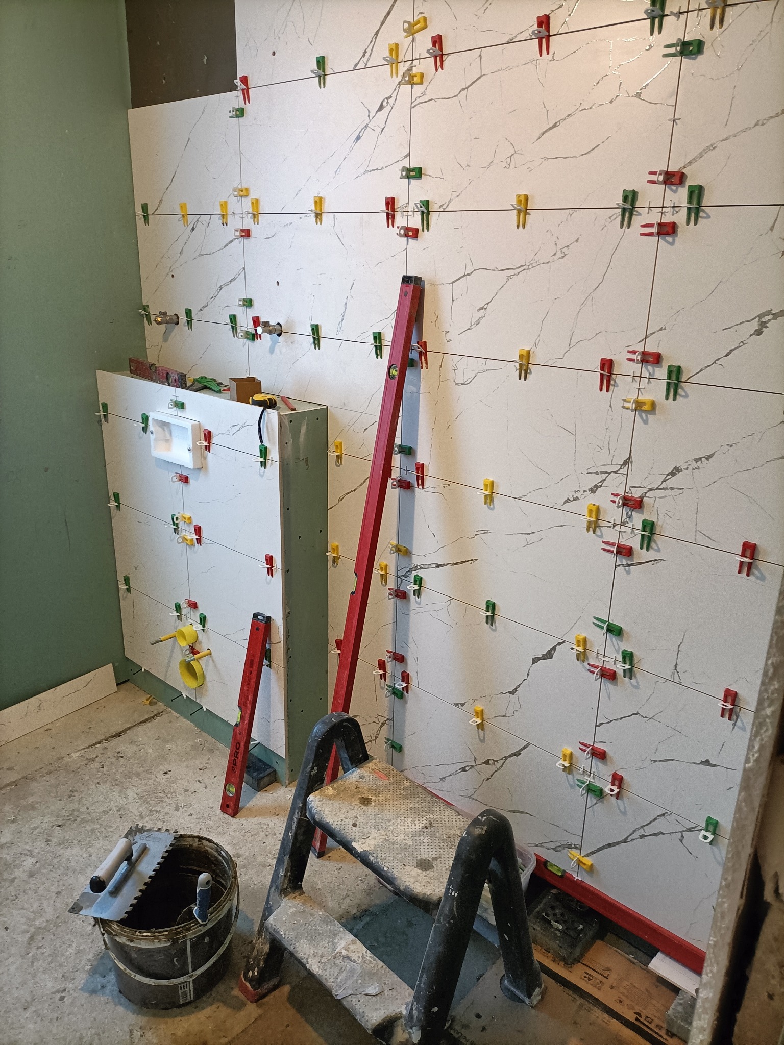 Montaż płytek imitujących marmur w łazience, z użyciem poziomicy, drabiny i systemu poziomowania płytek (klipsy w kolorach czerwonym, zielonym i żółtym).