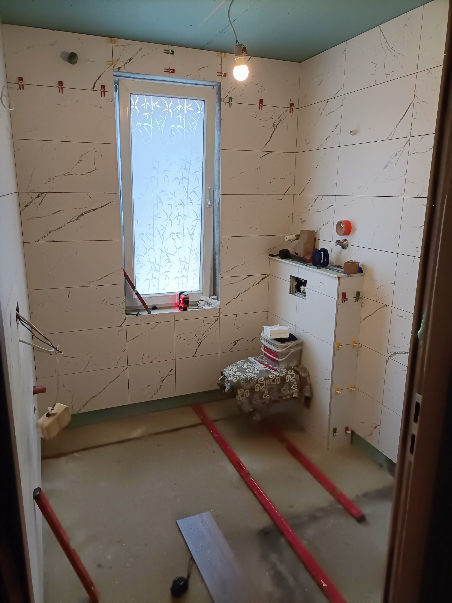 Łazienka w trakcie prac wykończeniowych, widoczne płytki imitujące marmur na ścianach, poziomowanie podłogi z użyciem listew, okno z dekoracyjną folią, wisząca żarówka i narzędzia na zabudowie...