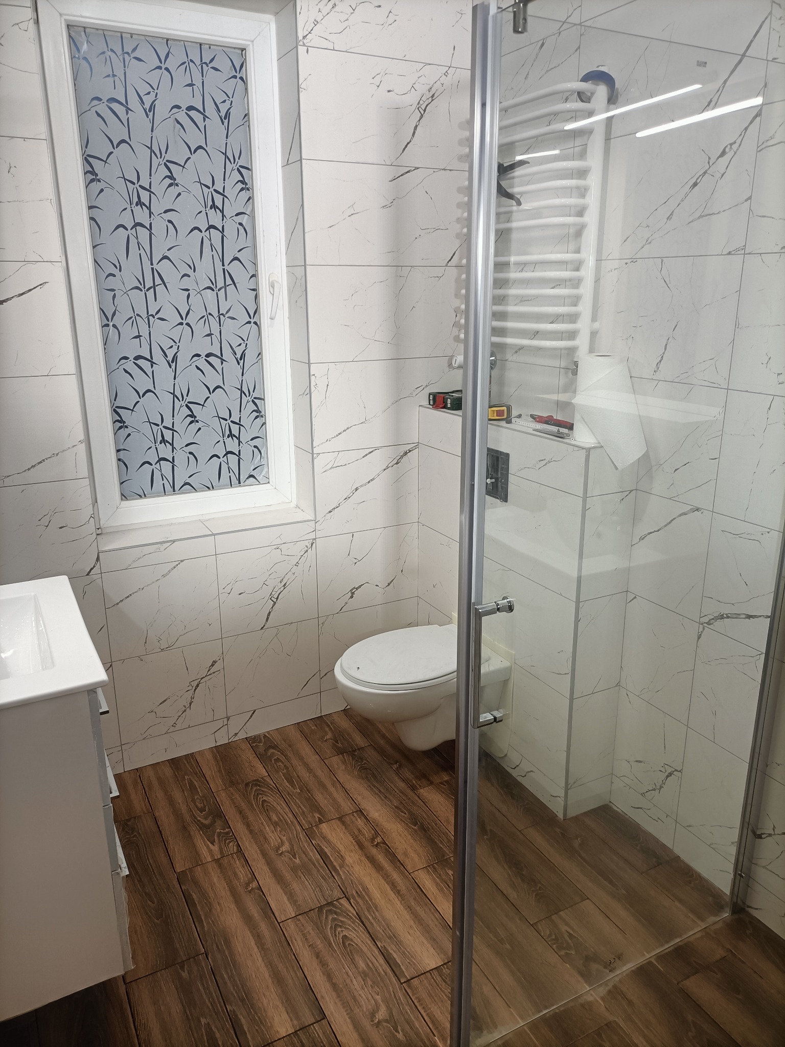 Nowoczesna łazienka po remoncie: białe marmurowe płytki ścienne, drewnopodobna podłoga, kabina prysznicowa ze szklanymi drzwiami, toaleta podwieszana i okno z folią imitującą bambus.