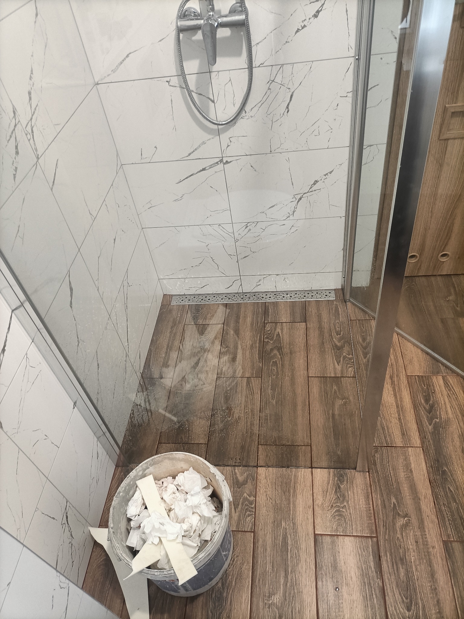 Ukończona kabina prysznicowa z odpływem liniowym, wyłożona płytkami imitującymi drewno na podłodze i białymi płytkami z szarymi żyłkami na ścianach, widoczne wiadro z zabrudzonymi materiałami po...