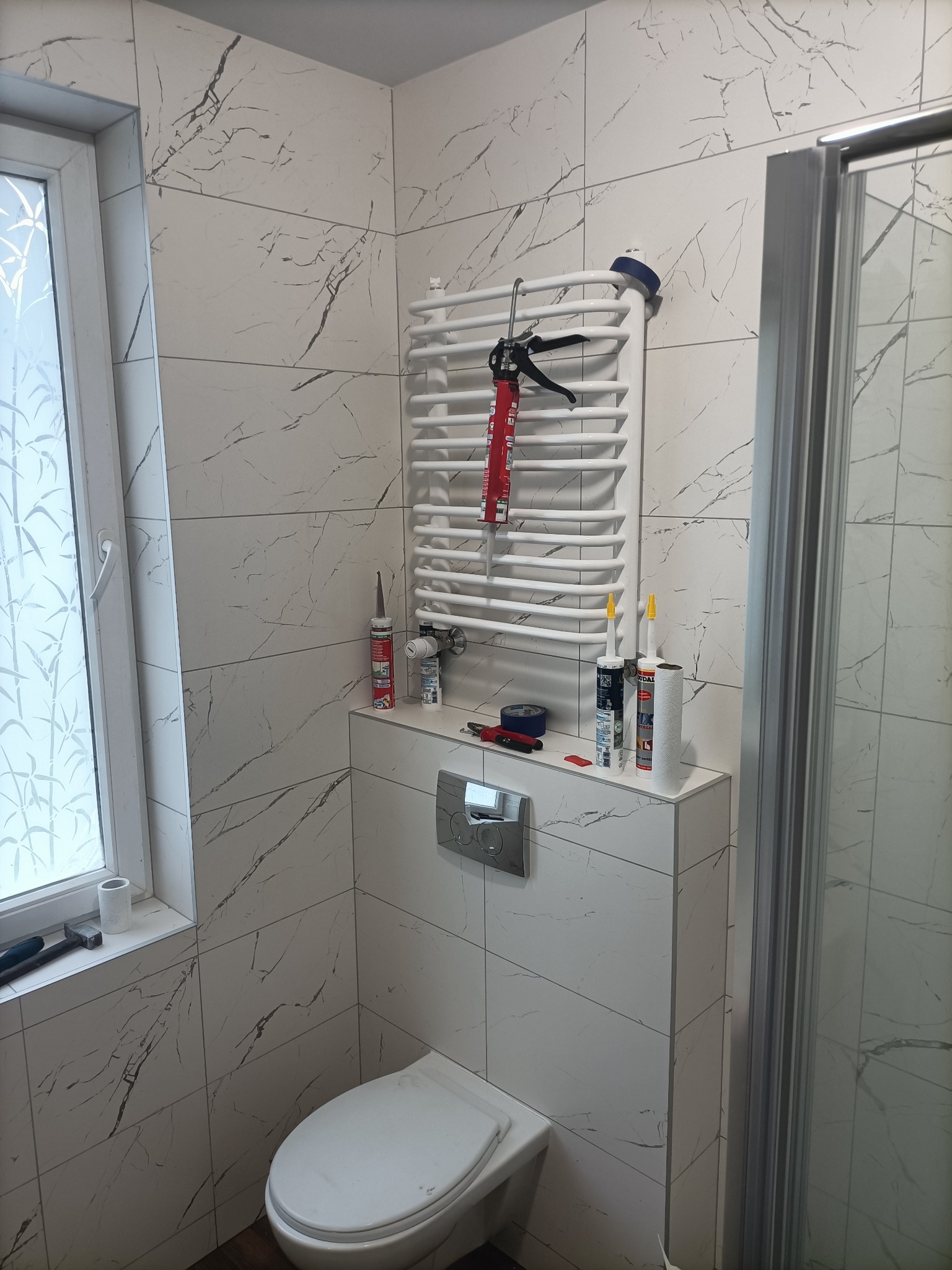 Biała łazienka w Werbkowicach z płytkami imitującymi marmur, z widoczną toaletą, grzejnikiem drabinkowym z aplikatorem silikonu, oraz akcesoriami do fugowania i silikonowania na zabudowie geberit.