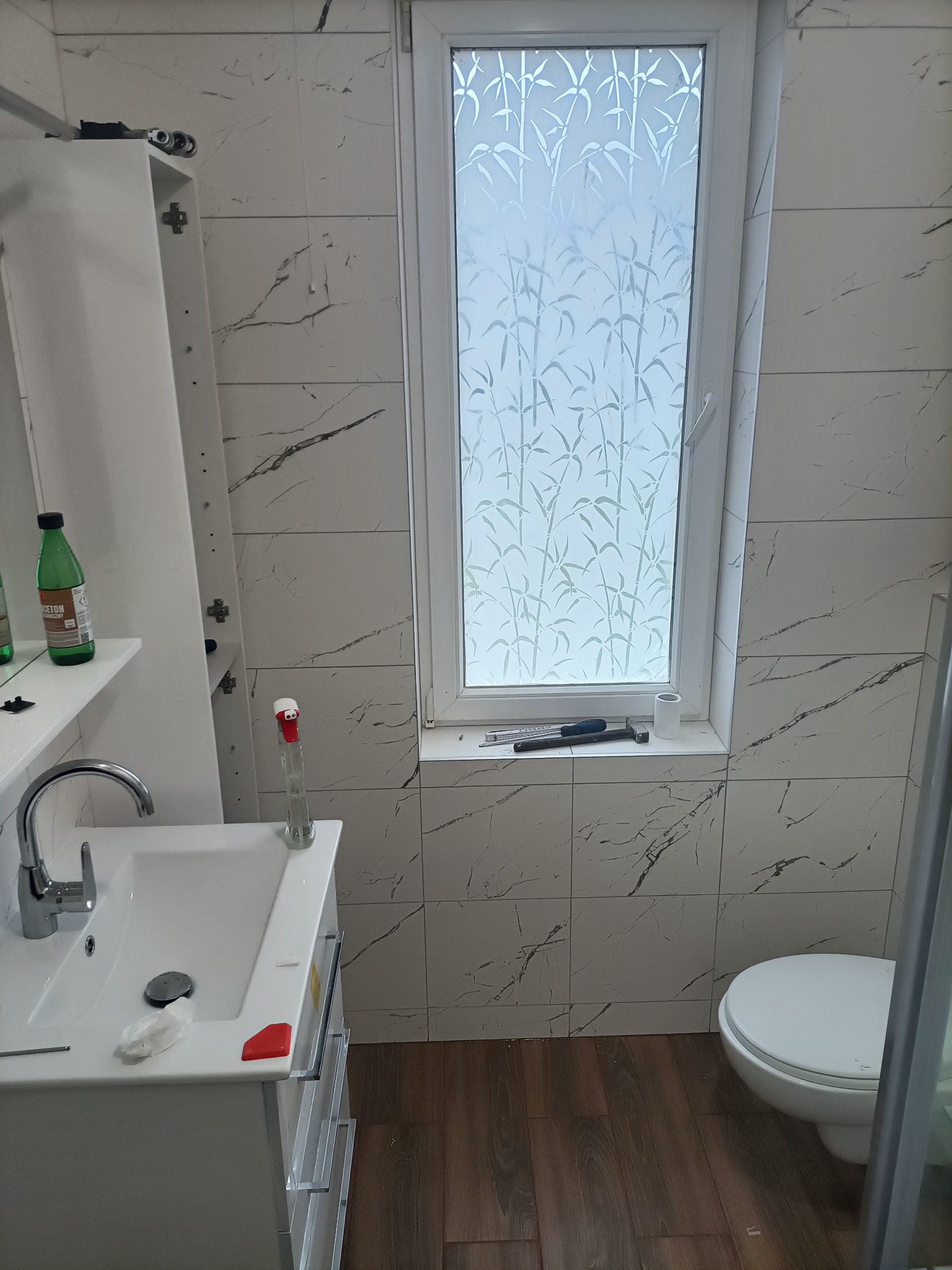 Łazienka w trakcie wykończenia: białe płytki imitujące marmur, umywalka z szafką, toaleta, okno z folią dekoracyjną, narzędzia na parapecie, niedokończona szafka wisząca.