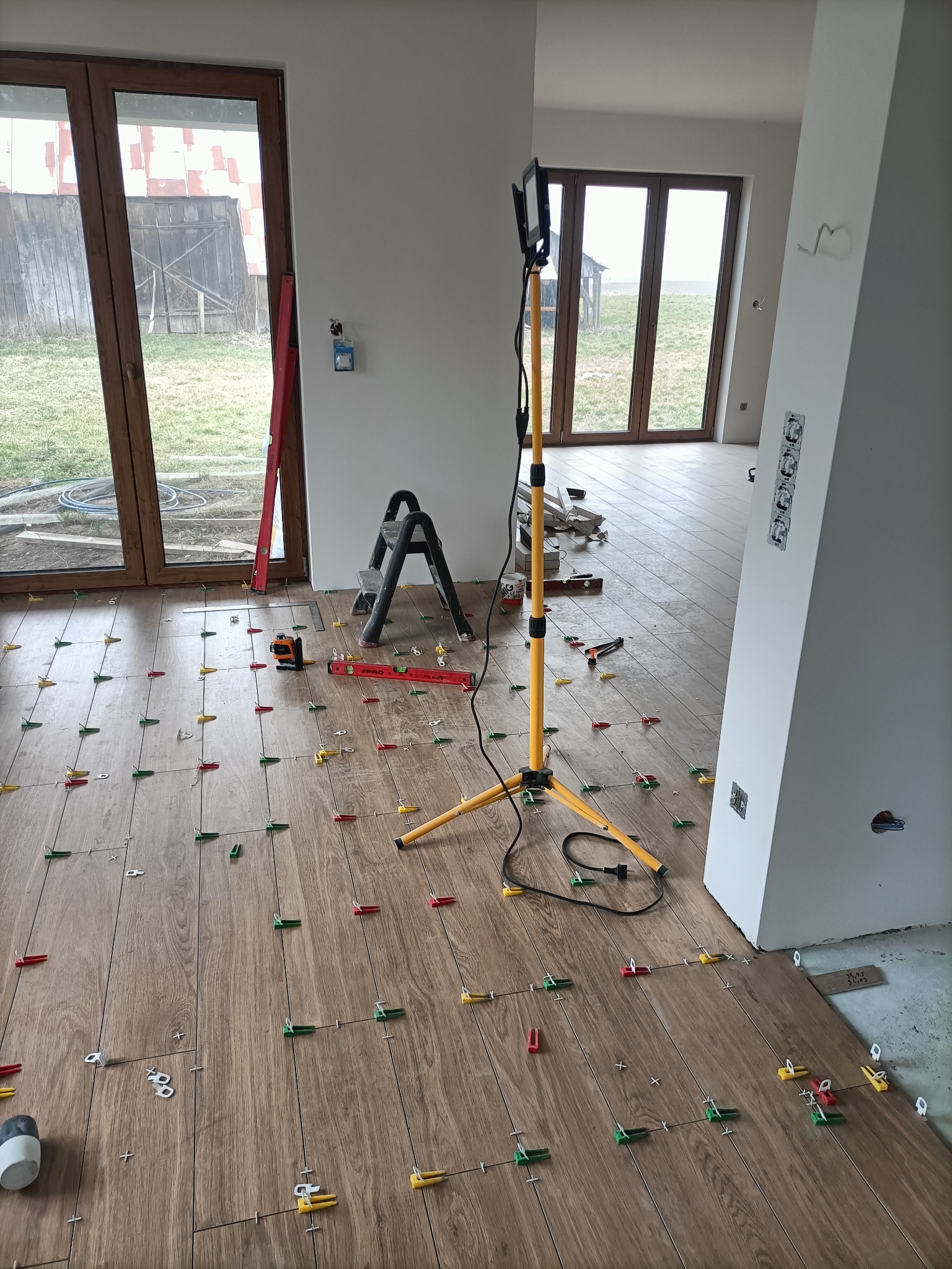 Układanie płytek podłogowych z użyciem klipsów poziomujących, widoczna drabina, poziomica laserowa i lampa budowlana w pomieszczeniu z dużymi oknami.