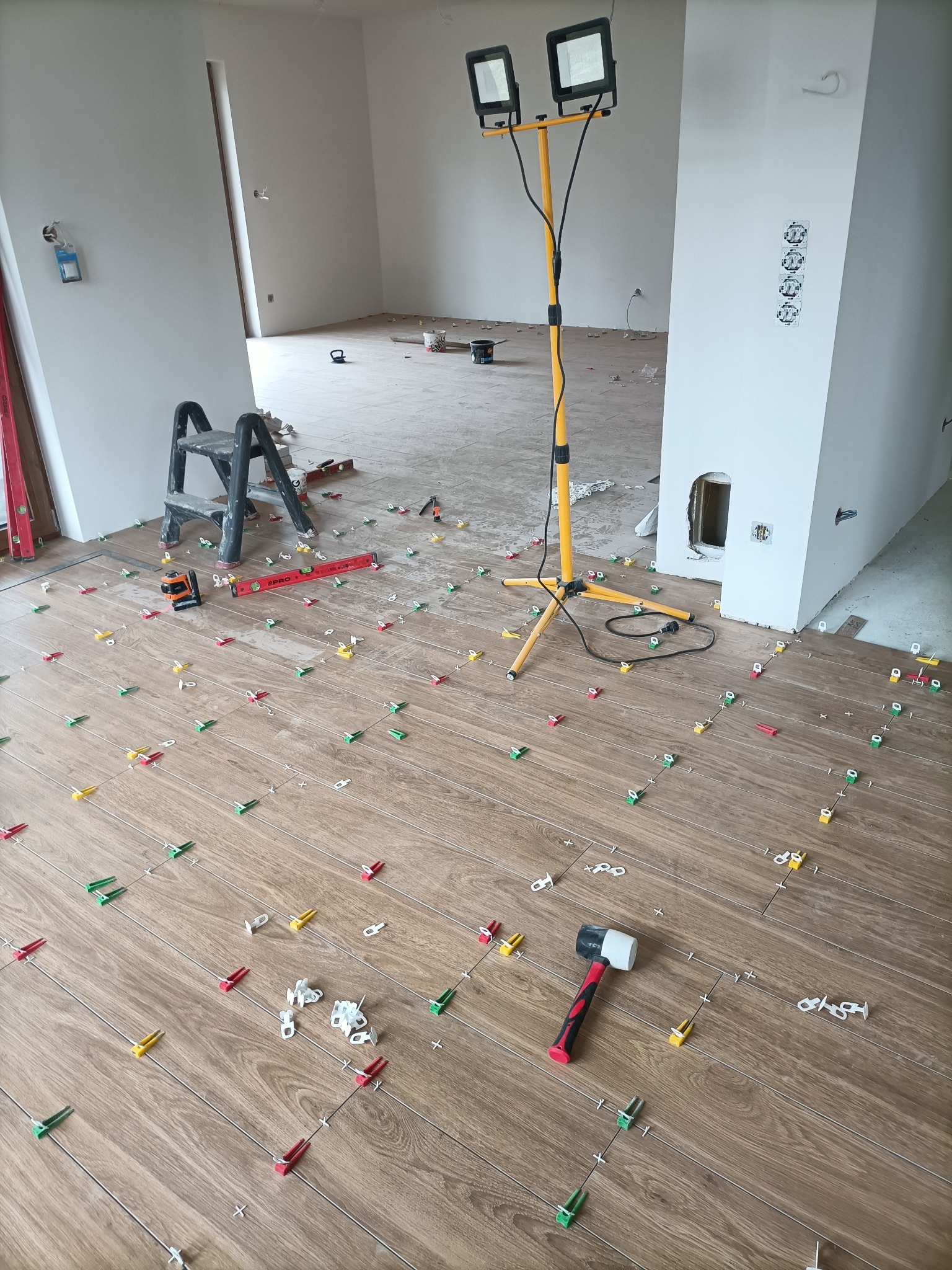 Układanie drewnopodobnych płytek podłogowych z użyciem poziomicy laserowej, gumowego młotka i klipsów poziomujących; widoczna drabinka i oświetlenie budowlane.