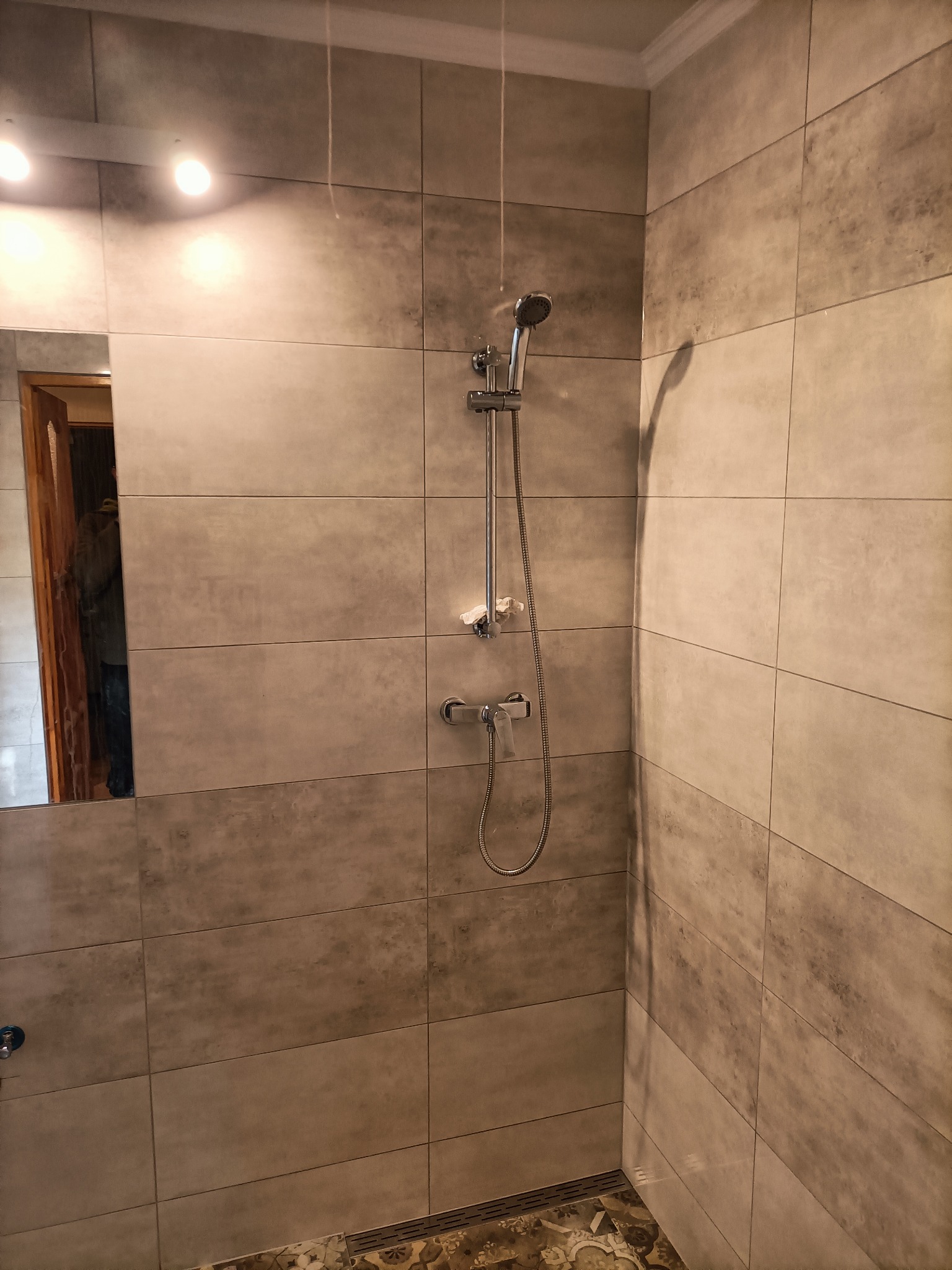 Wykończona łazienka z szarymi, prostokątnymi płytkami imitującymi beton, chromowaną armaturą prysznicową i fragmentem dekoracyjnej podłogi w stylu vintage.