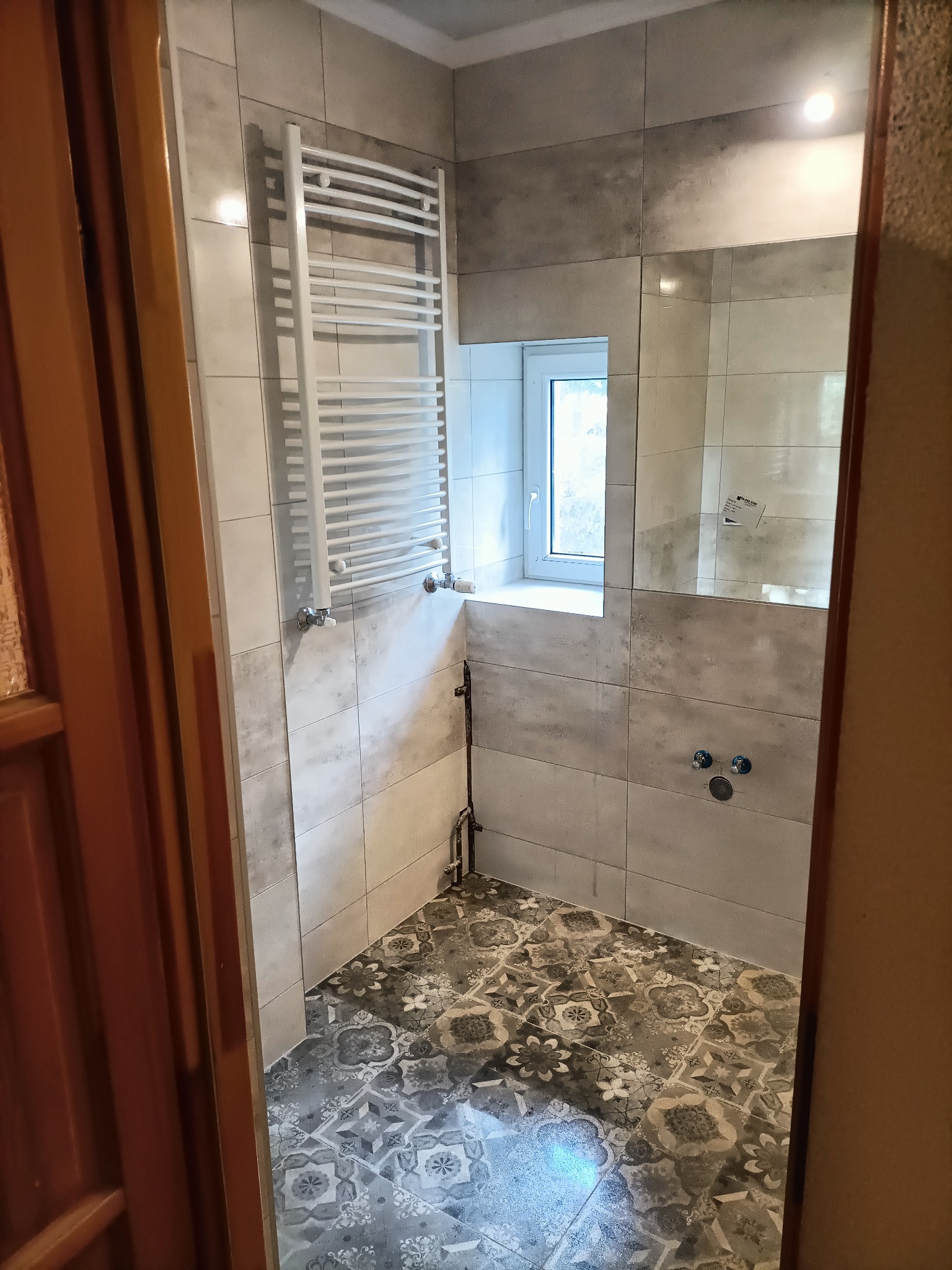 Wykończona łazienka z szaro-białymi płytkami na ścianach i dekoracyjną, wzorzystą podłogą, widoczny grzejnik drabinkowy, okno i instalacja wodna bez armatury.