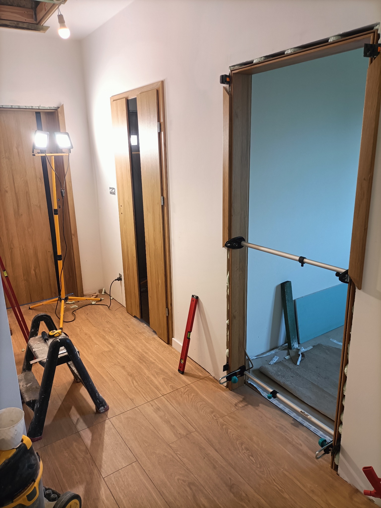 Montaż ościeżnicy w wyremontowanym wnętrzu, widoczne drewniane drzwi, poziomica, drabina i lampa budowlana