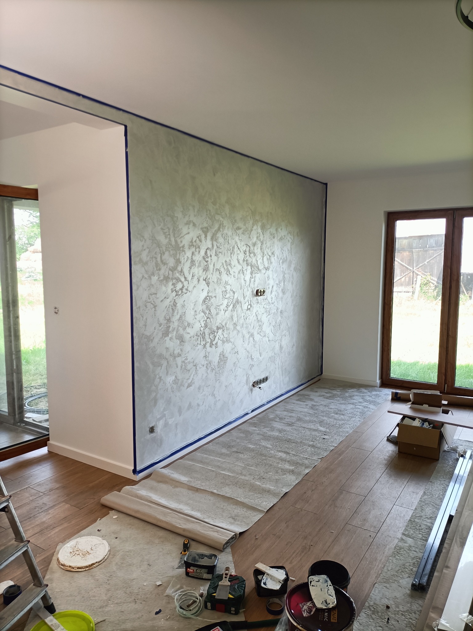 Ściana w trakcie dekoracyjnego wykończenia z efektem strukturalnym, zabezpieczona taśmą malarską, podłoga częściowo zakryta folią ochronną, widoczne narzędzia i materiały malarskie, przygotowane...