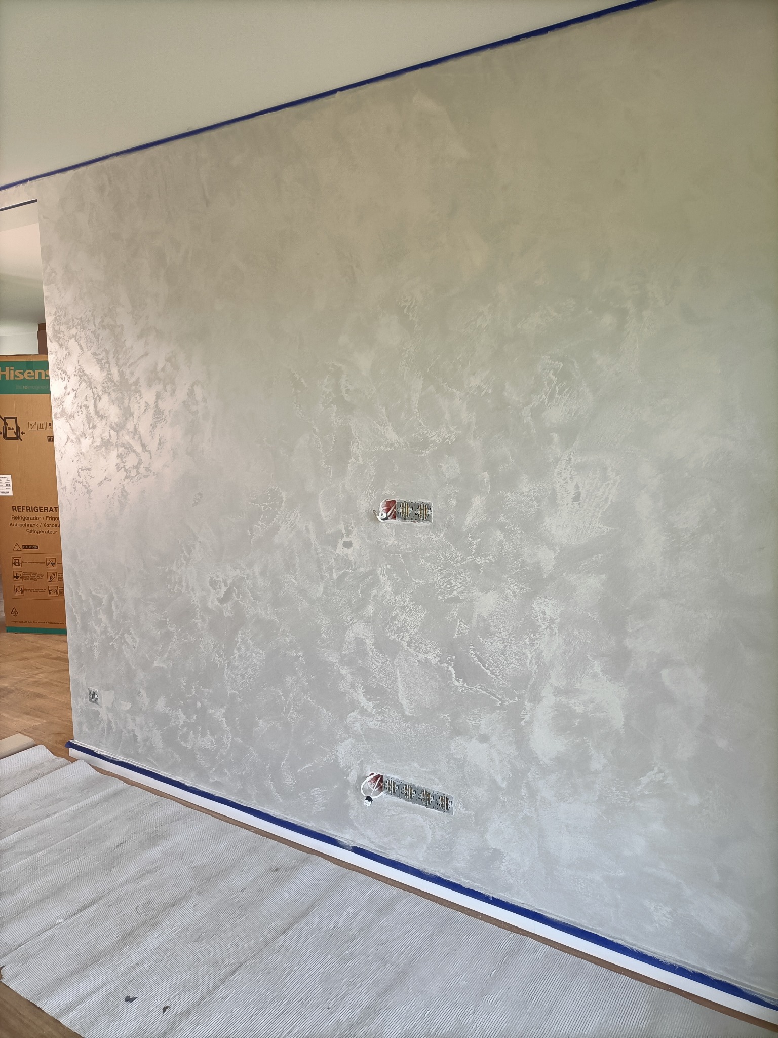 Ściana pokryta dekoracyjną, jasną gładzią w odcieniach szarości i bieli, zabezpieczona niebieską taśmą malarską przy suficie i podłodze, z widocznymi gniazdkami elektrycznymi.