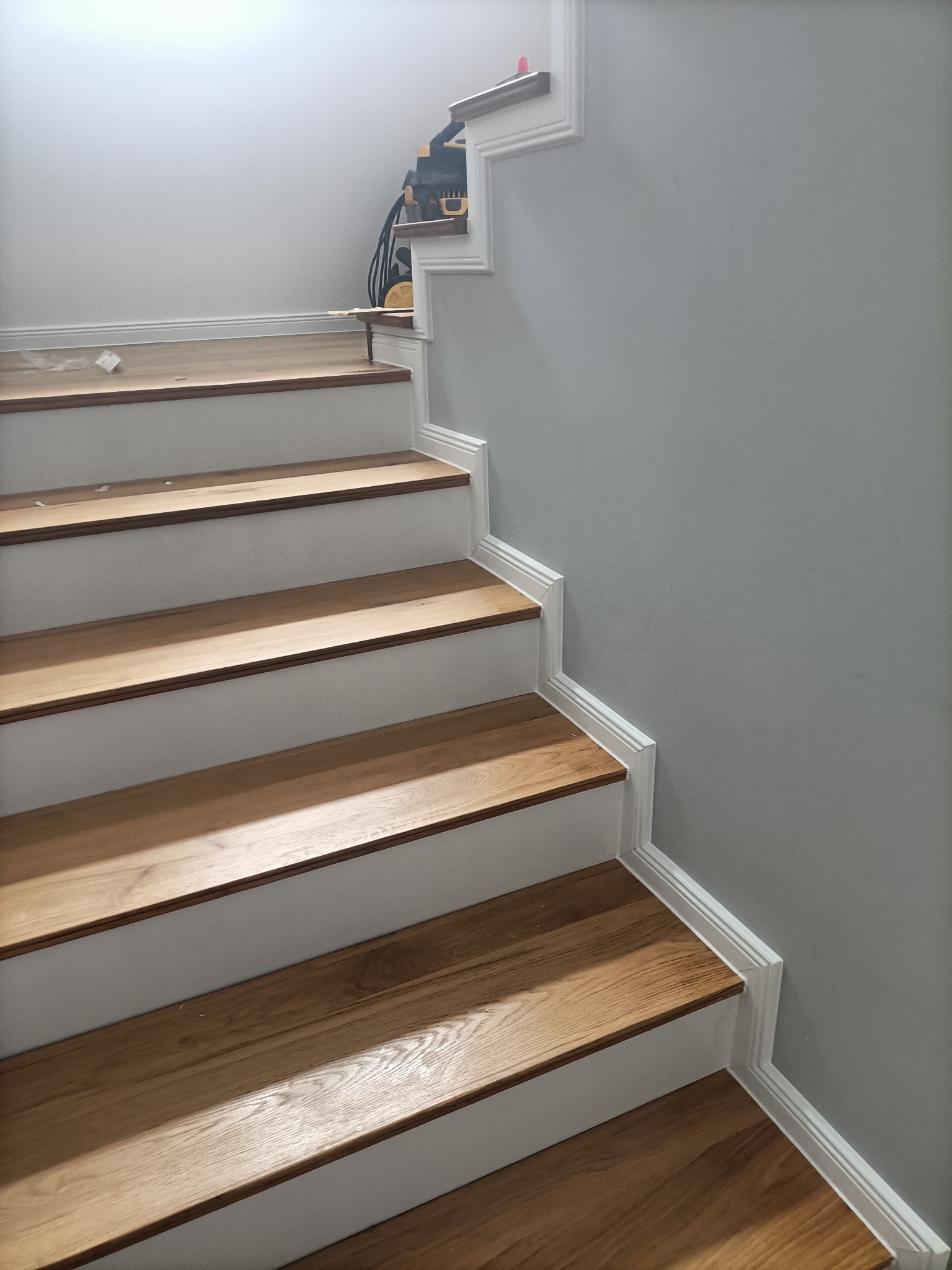 Drewniane schody z białymi podstopnicami i listwami przypodłogowymi, częściowo wykończone, z odkurzaczem widocznym na górnym biegu.