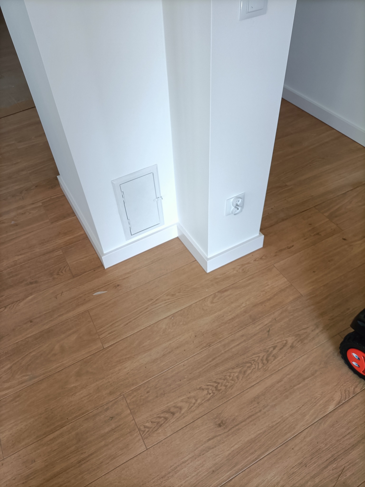 Świeżo pomalowany narożnik ściany w mieszkaniu z widoczną klapką rewizyjną i gniazdkiem elektrycznym, wykończony przypodłogówką, na podłodze panele imitujące drewno.