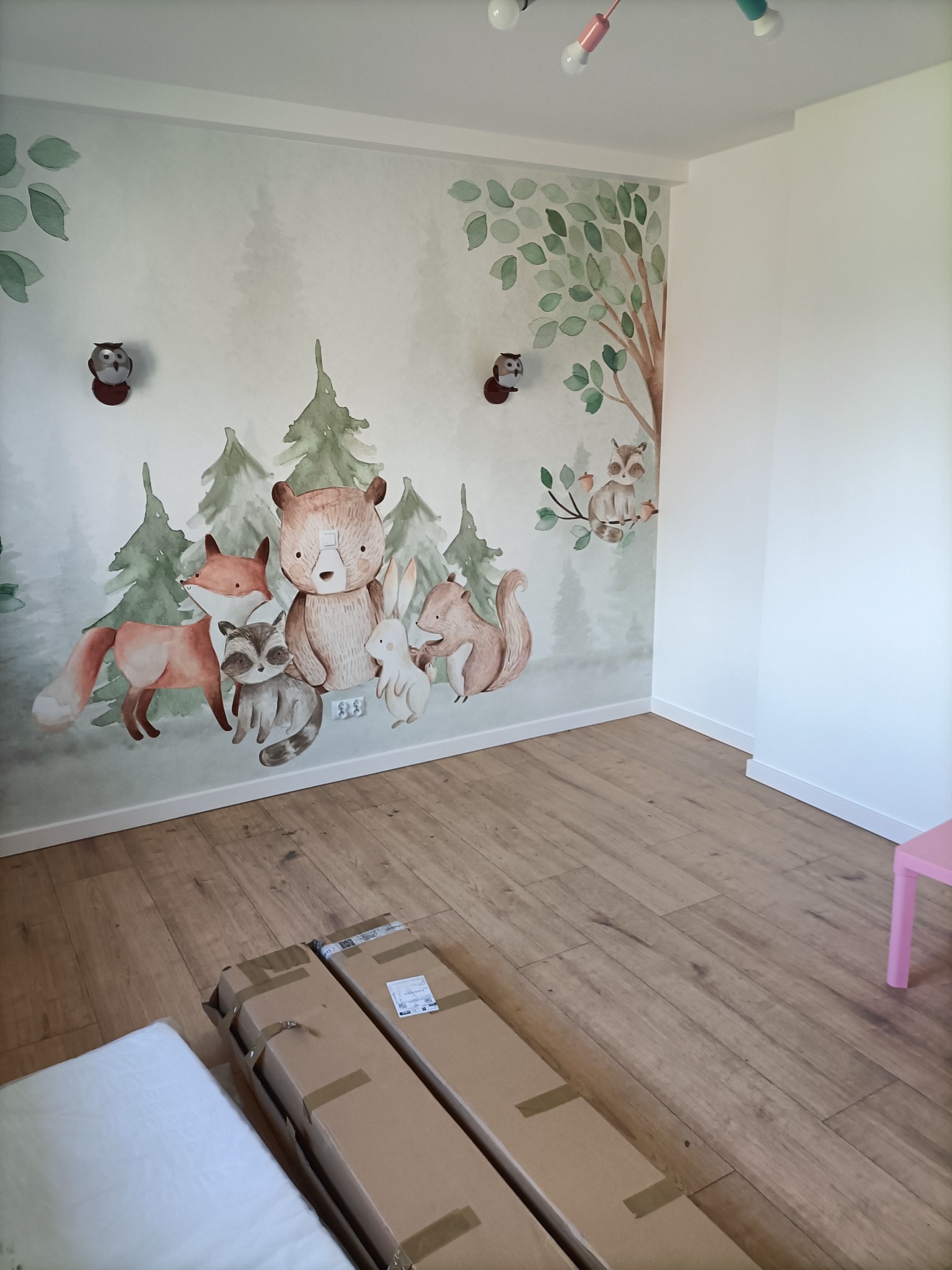 Pokój dziecięcy z ręcznie malowaną ścianą przedstawiającą leśne zwierzęta, drewniana podłoga i zapakowane elementy wyposażenia, widoczne lampki w pastelowych kolorach.