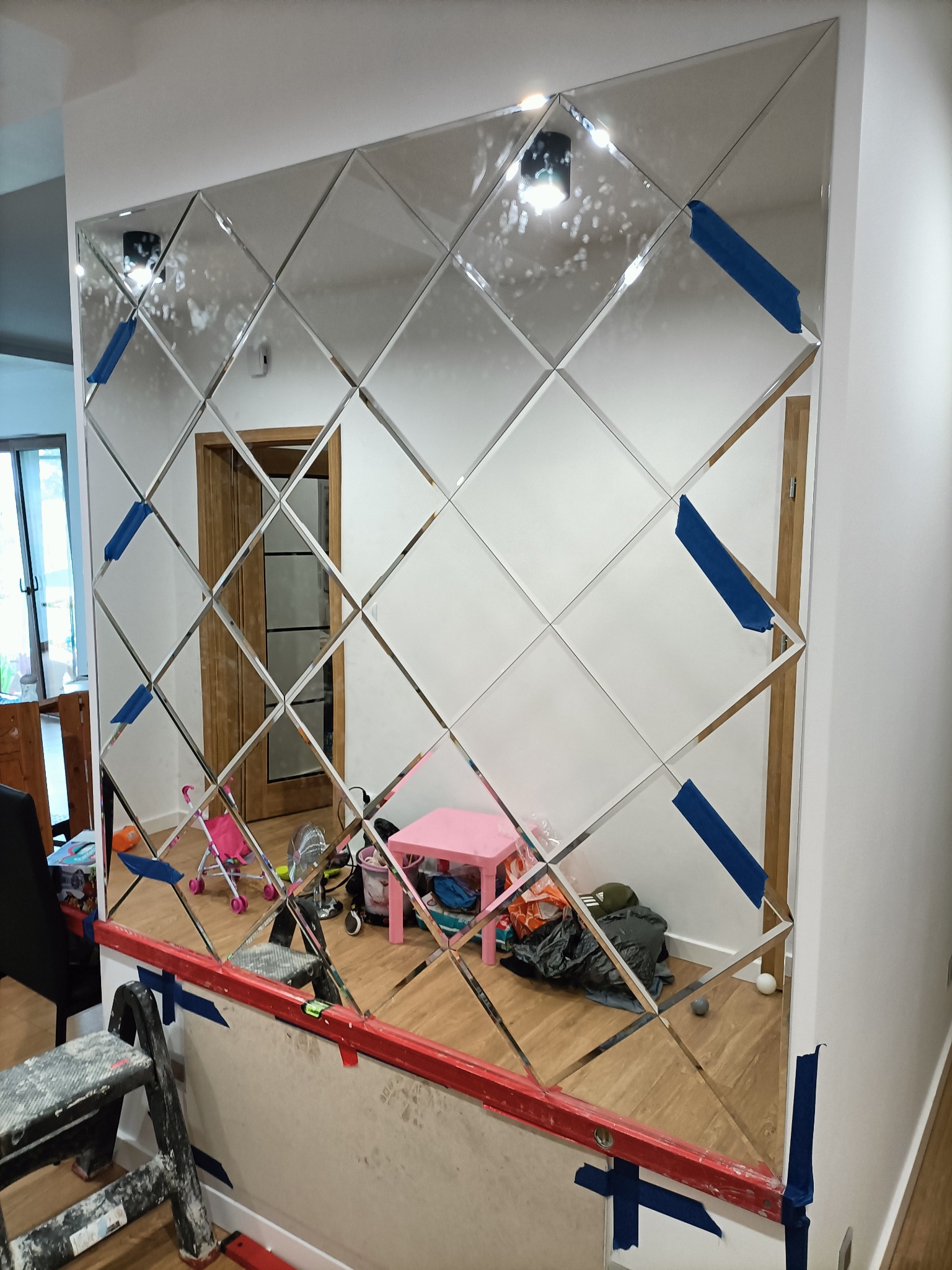 Montaż lustrzanej ściany z romboidalnych elementów, zabezpieczonych niebieską taśmą malarską, widoczna poziomica i drabina, w tle pokój dziecięcy.