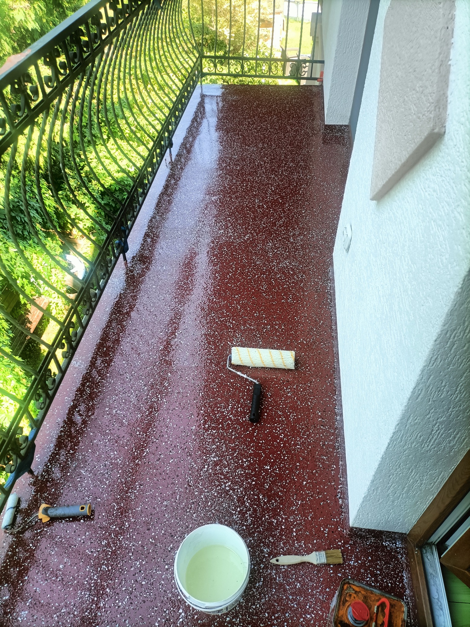 Świeżo pomalowany balkon z dekoracyjną, bordową posadzką w białe plamki, widoczne narzędzia malarskie: wałek, pędzel, kuweta i wiadro z farbą w rogu, metalowa balustrada w tle.