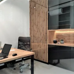 Nowoczesne biuro z drewnianym biurkiem, czarnym krzesłem obrotowym i panelem dekoracyjnym z pionowych listew, wyposażone w laptop, smartfon, mysz bezprzewodową i szklane butelki z wodą.