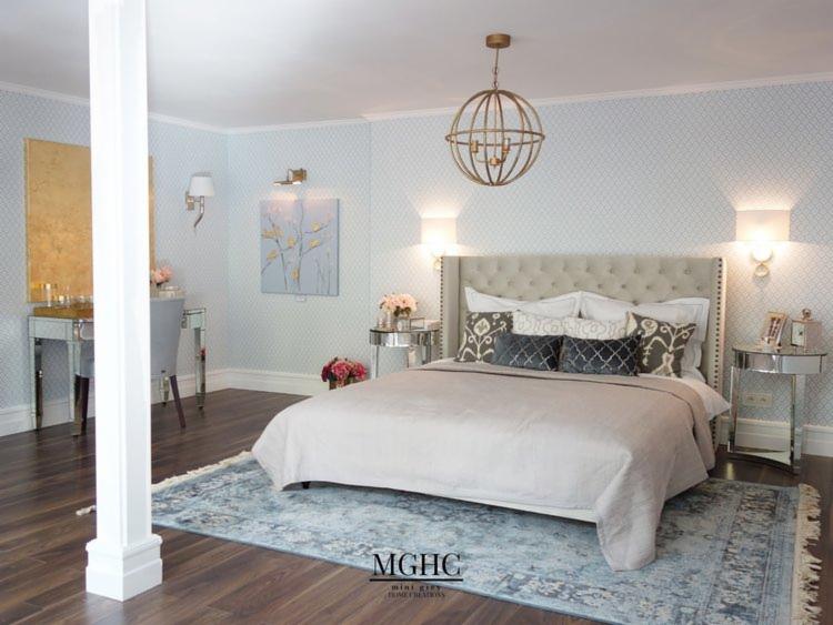 Elegancka sypialnia z tapetą w delikatny wzór, łóżkiem z tapicerowanym zagłówkiem i lustrzanymi stolikami nocnymi, oświetlona kulistym żyrandolem i kinkietami, z dywanem w odcieniach błękitu...