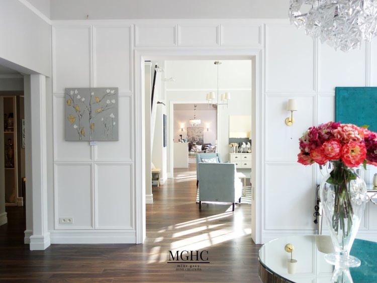 Eleganckie wnętrze z białymi panelami ściennymi, ciemną drewnianą podłogą i widokiem na dalsze pokoje, oświetlone naturalnym światłem wpadającym przez drzwi. Na ścianie obraz z motywem drzewa, a...