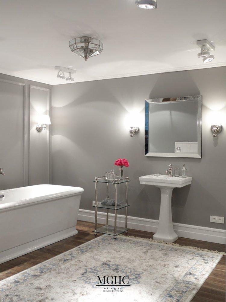 Elegancka łazienka w odcieniach szarości z wolnostojącą wanną, umywalką na postumencie, lustrem w ramie, kryształowym żyrandolem i dywanem w stylu vintage.