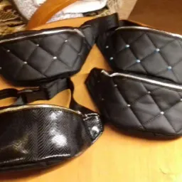 Cztery czarne saszetki biodrowe, w tym trzy pikowane z ozdobnymi dżetami i jedna z fakturą włókna węglowego, leżące na drewnianym stole.