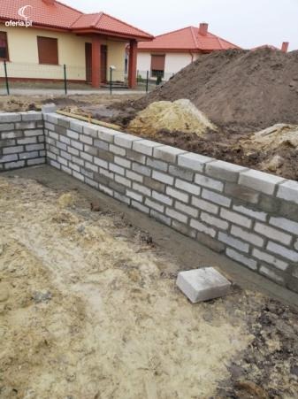 Wznoszenie muru fundamentowego z białej cegły silikatowej, widoczne świeże spoiny. W tle kopiec ziemi i dom jednorodzinny z czerwoną dachówką.