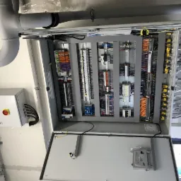 Otwarta szafa sterownicza z bogatym okablowaniem, widoczne liczne złączki, kable w różnych kolorach i przekrojach, oraz dodatkowa puszka sterownicza z czerwonym przyciskiem bezpieczeństwa...