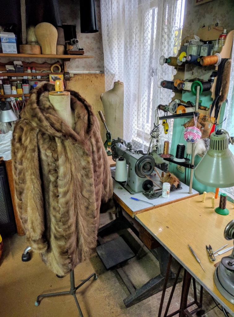 Futro z kapturem na manekinie w pracowni, w tle maszyna do szycia, nici i narzędzia.