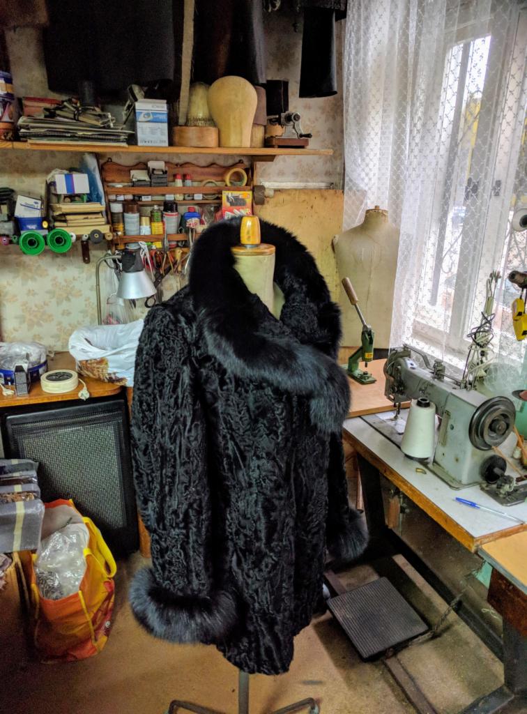 Gotowe, czarne futro astrachańskie z futrzanym kołnierzem i mankietami, prezentowane na manekinie w pracowni krawieckiej z maszyną do szycia i półkami z narzędziami w tle.