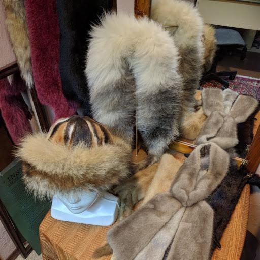 Ekspozycja futrzanych akcesoriów: czapka z futrzanym otokiem na białym manekinie głowy, szale w odcieniach bieli, szarości i brązu, wiszące na wieszakach i ułożone na stole.