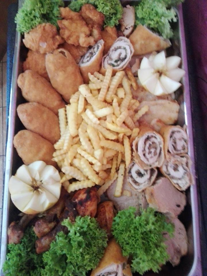 Metalowa taca z różnorodnym cateringiem: roladki mięsne z ciemnym farszem, kotlety w panierce, frytki, pieczone mięso, ozdobiona sałatą i jabłkami w kształcie kwiatów.