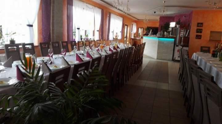 Ustawione stoły z białymi obrusami, bordowymi serwetkami i szklankami w przestronnej sali z pomarańczowymi ścianami, widoczne także rośliny doniczkowe.